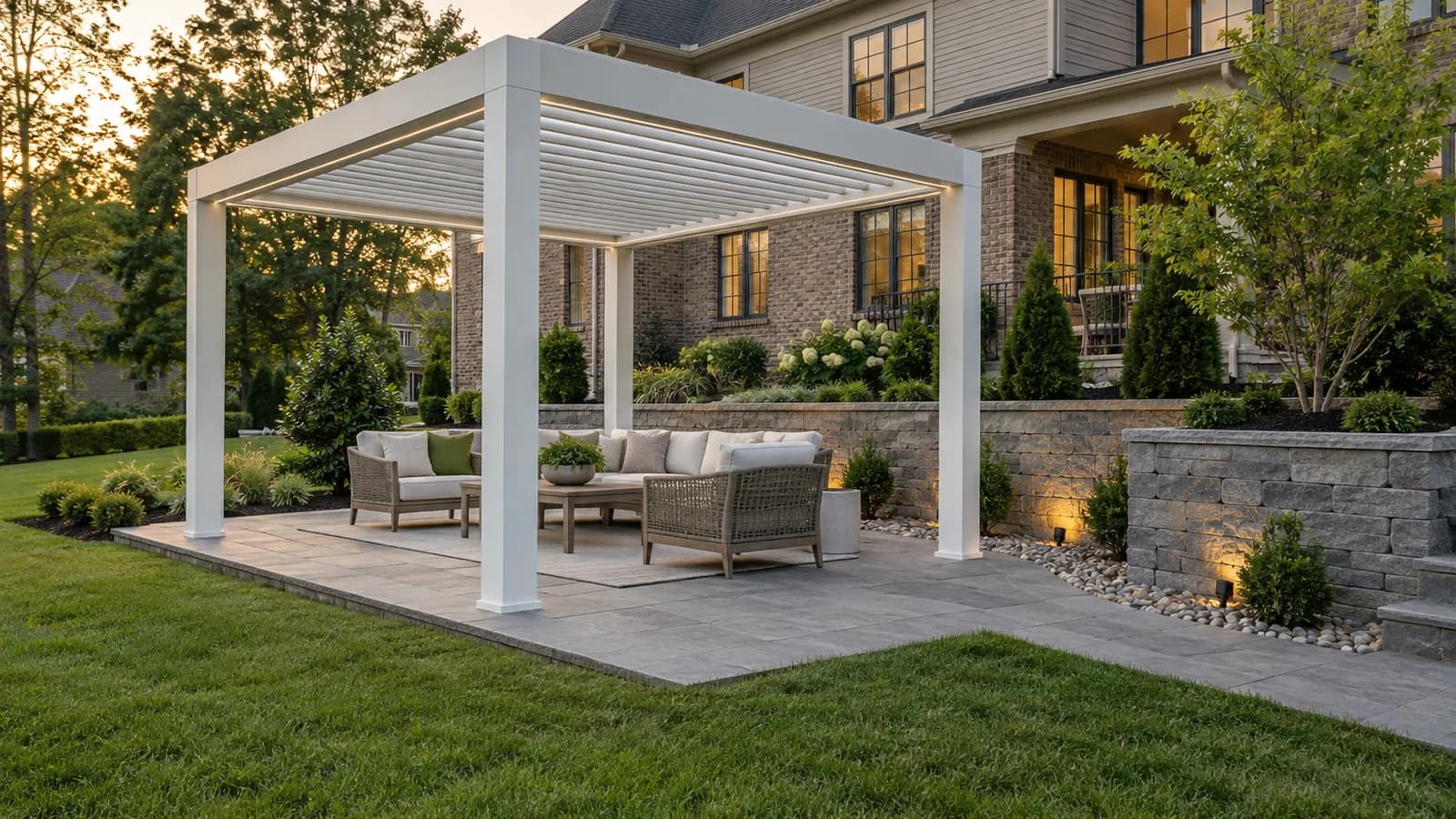 White aluminum louvered pergola on a stone patio beside a low gray retaining wall and landscaped slope